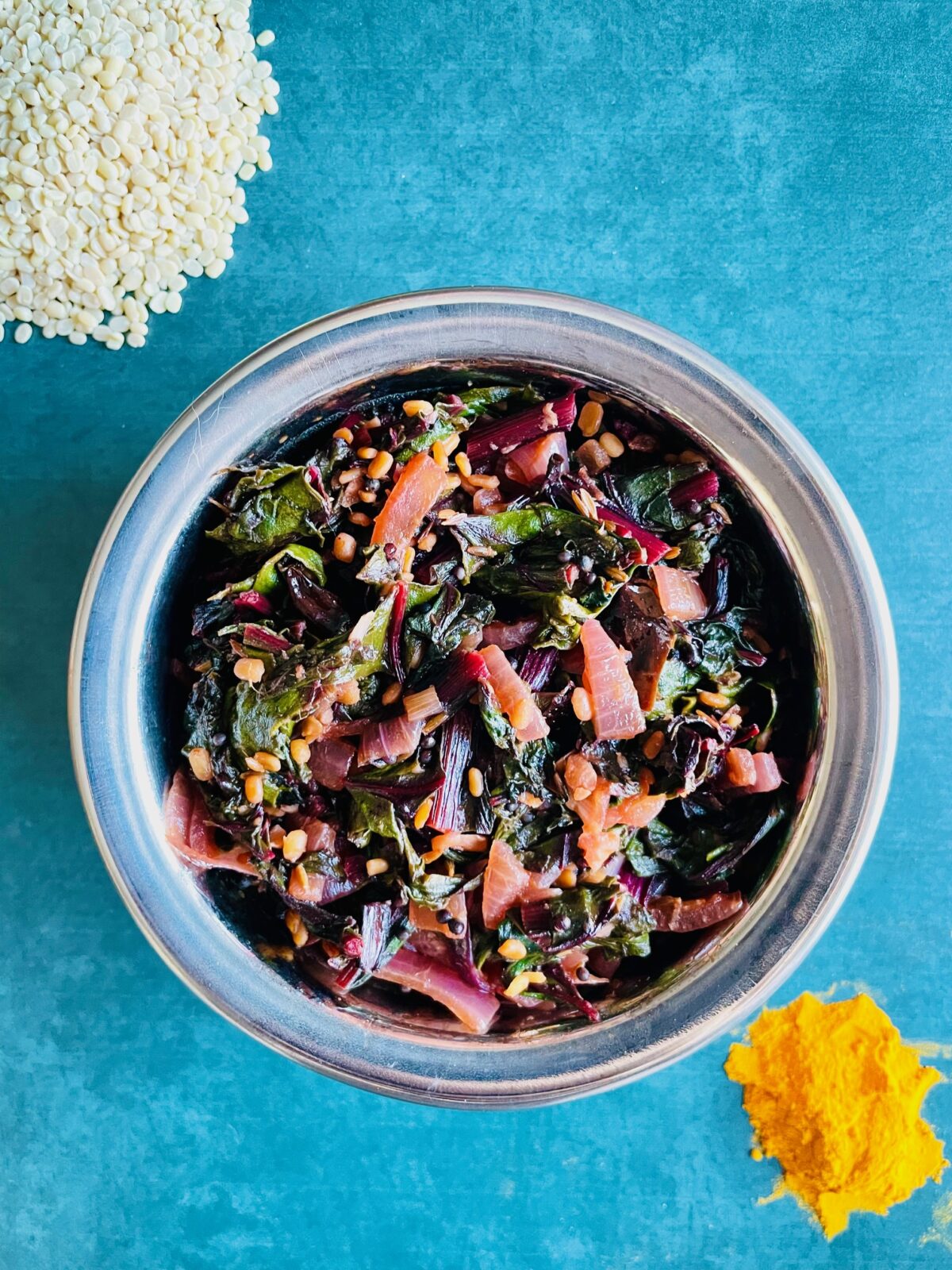 Indian sauteed beet greens in copper bowl, turmeric powder in front and urad dal in the back.