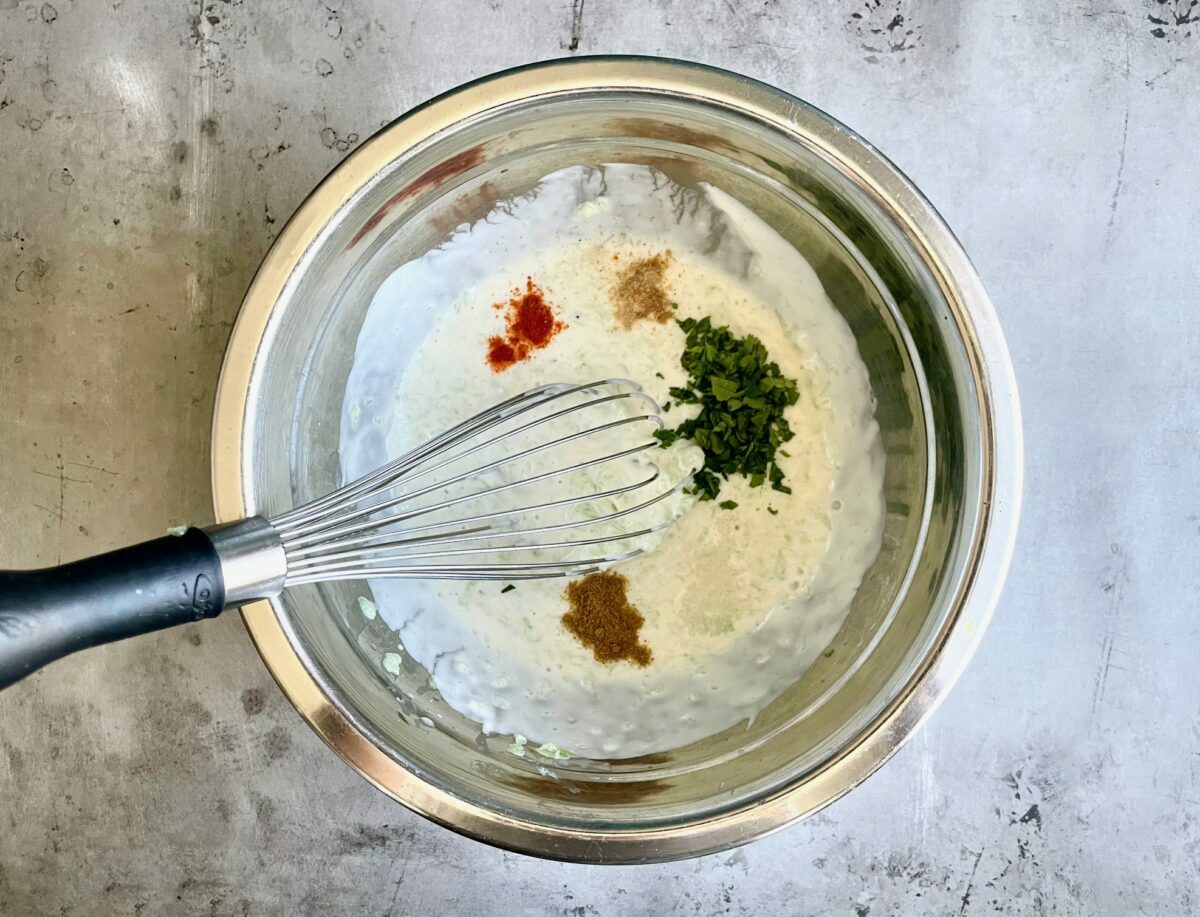 Cilantro, mint and spices on top of yogurt in a stainless steel bowl. Whisk inside the bowl.