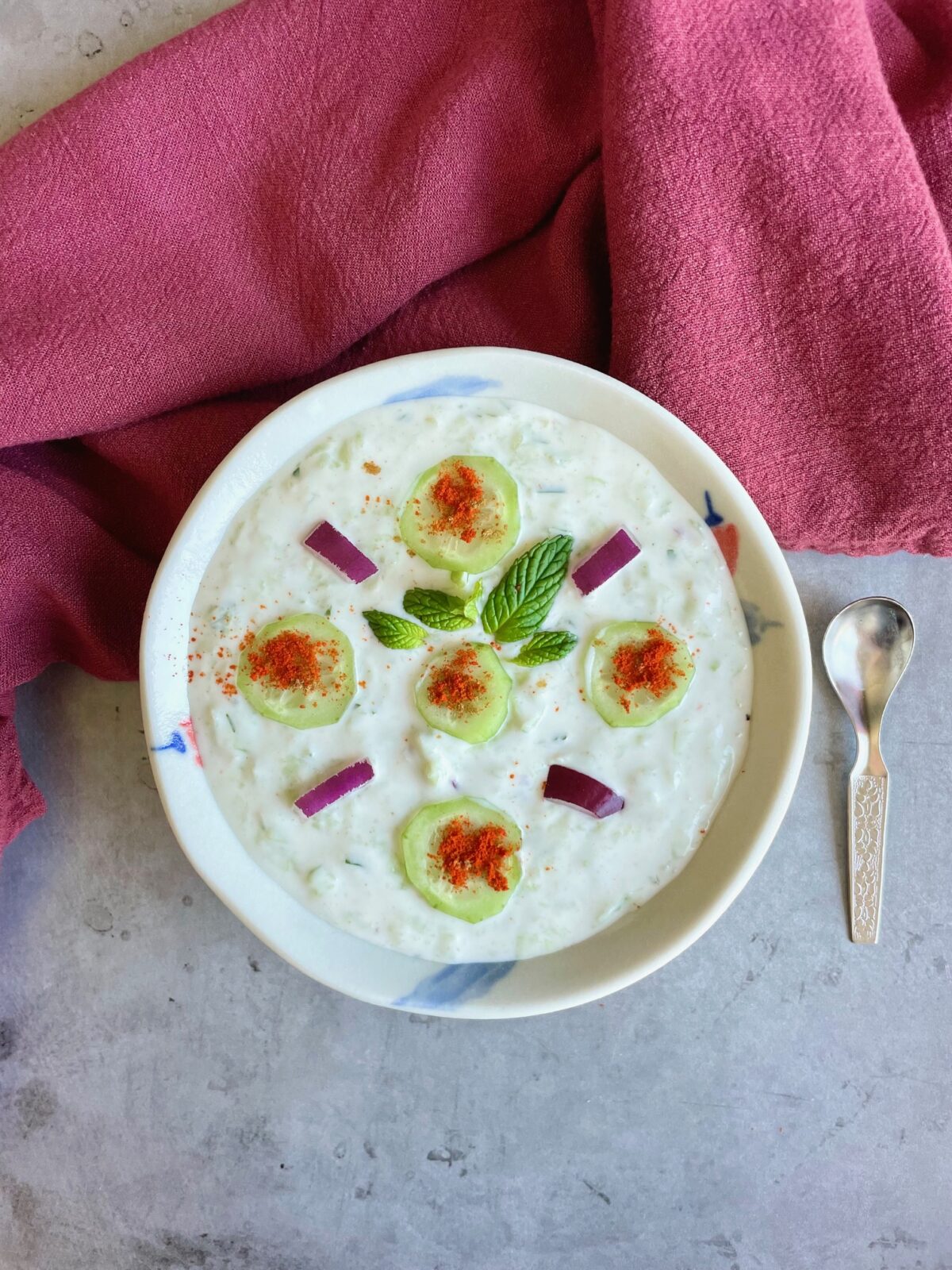 Cucumber raita recipe for biryani with fresh cucumbers, mint and red onion slices. Stainless steel spoon next to the bowl. Red towel underneath serving bowl.