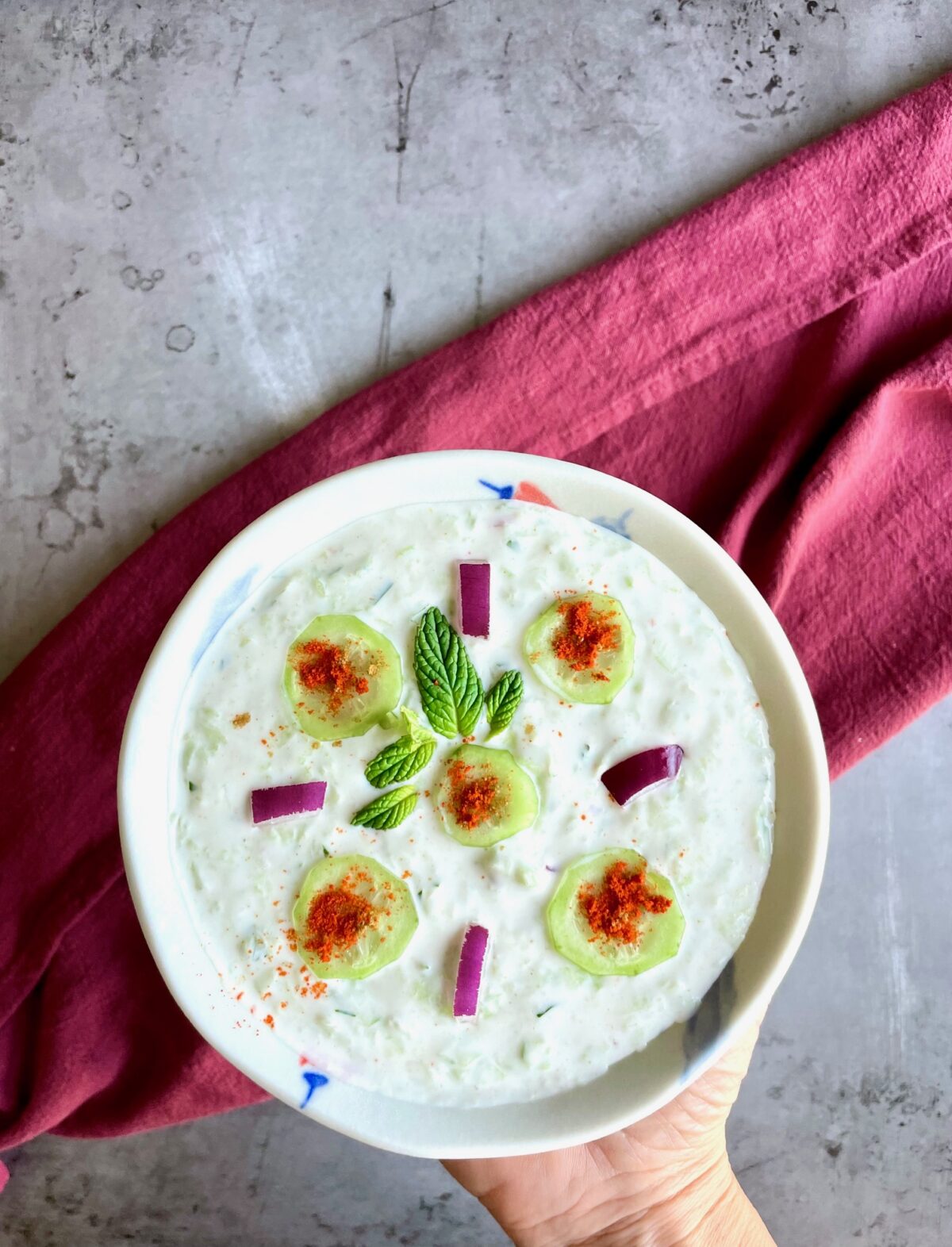 Cucumber raita with red onion, mint leaves and cucumber slices in a bowl. Bowl is being held by a hand.