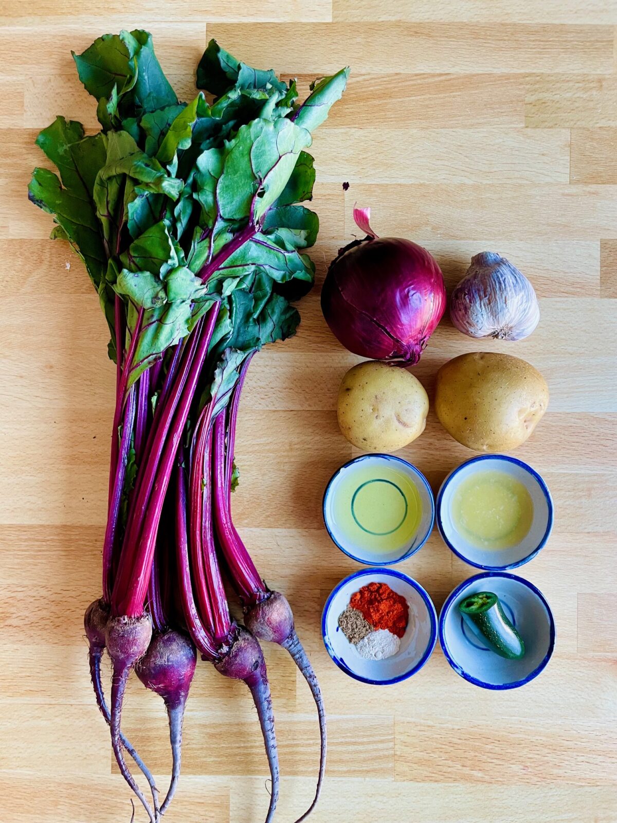 Ingredients for Ethiopian Beet Recipe: whole baby beets with greens attached, whole red onion, purple garlic clove, yellow potatoes, and ingredients in bowls: vegetable oil, fresh lemon juice, spices and half a jalapeno.