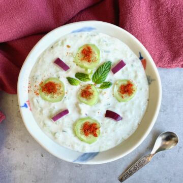 Raita with fresh cucumbers, mint and red onion slices. Stainless steel spoon next to the bowl. Red towel underneath serving bowl.
