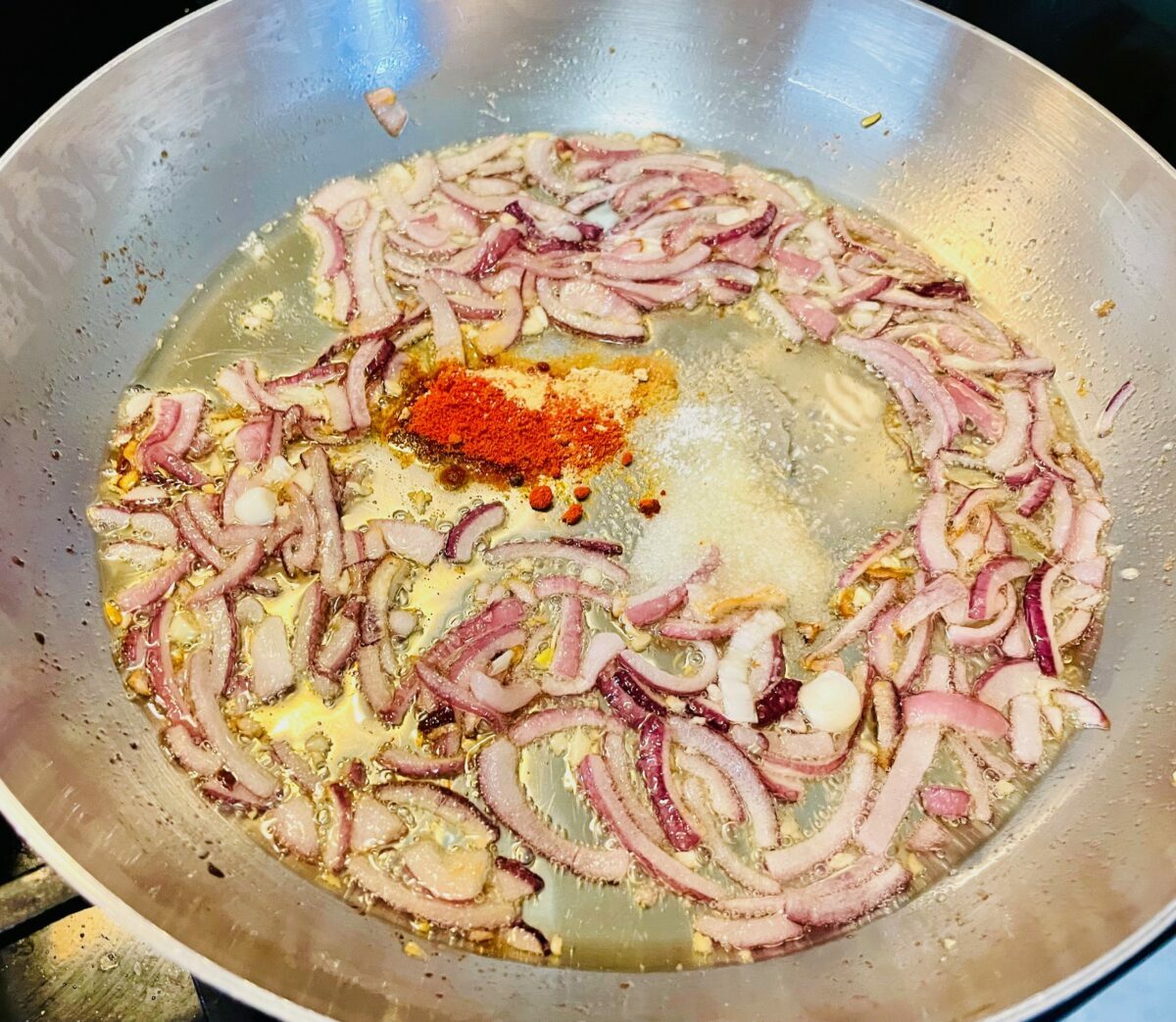 Sliced red onion with spices cooking in oil in a stainless steel pan.