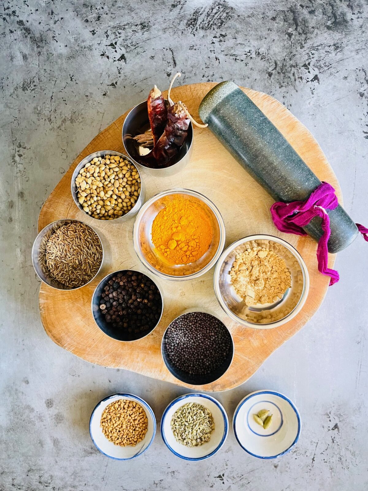 Whole spices for Indian curry powder in bowls: whole red chilies, coriander seeds, cumin seeds, black peppercorn, turmeric powder, ginger powder, mustard seeds, fenugreek seeds, fennel seeds and cardamom pods.
