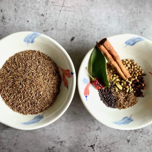 Cumin seeds in bowl and whole spices for curry powder in bowl. Whole spices: cinnamon stick, cumin seeds, curry leaf, coriander seeds, brown mustard seeds, fenugreek seeds, red chili, cardamom pods and whole cloves.
