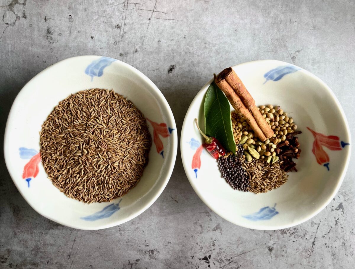 Cumin seeds in bowl and whole spices for curry powder in bowl. Whole spices: cinnamon stick, cumin seeds, curry leaf, coriander seeds, brown mustard seeds, fenugreek seeds, red chili, cardamom pods and whole cloves.