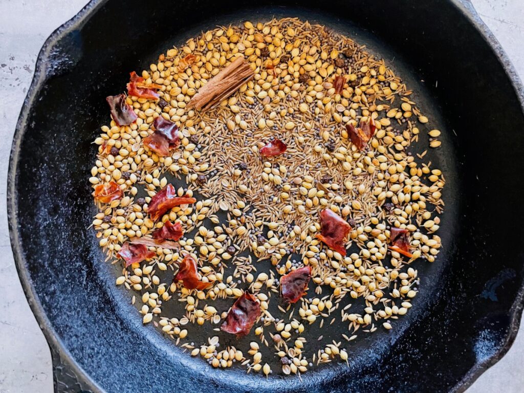Whole spices for masala mix in a cast iron pan: coriander seeds, cumin seeds, brown mustard seeds, Ceylon cinnamon, red chilies, and black peppercorn.