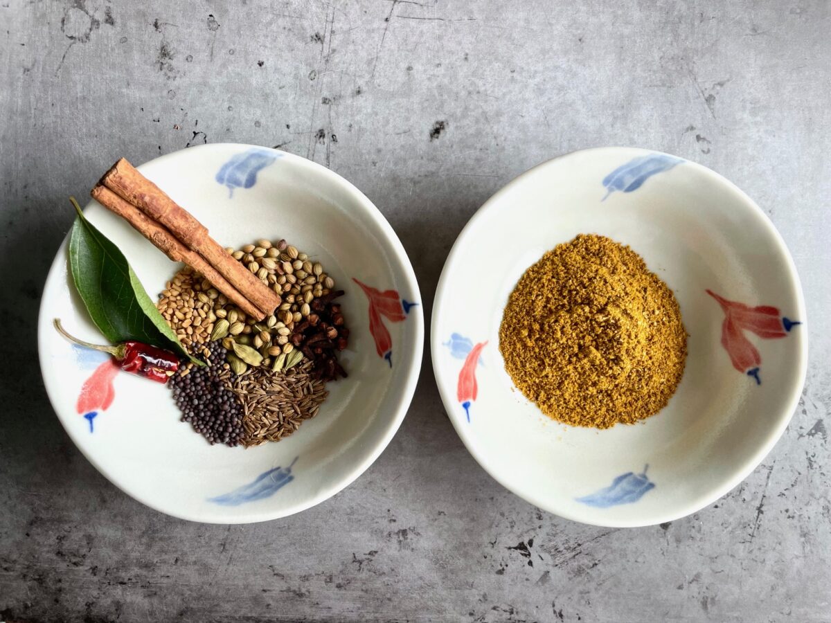 Whole spices for curry powder in bowl and curry powder in bowl. Whole spices: cinnamon stick, cumin seeds, curry leaf, coriander seeds, brown mustard seeds, fenugreek seeds, red chili, cardamom pods and whole cloves.
