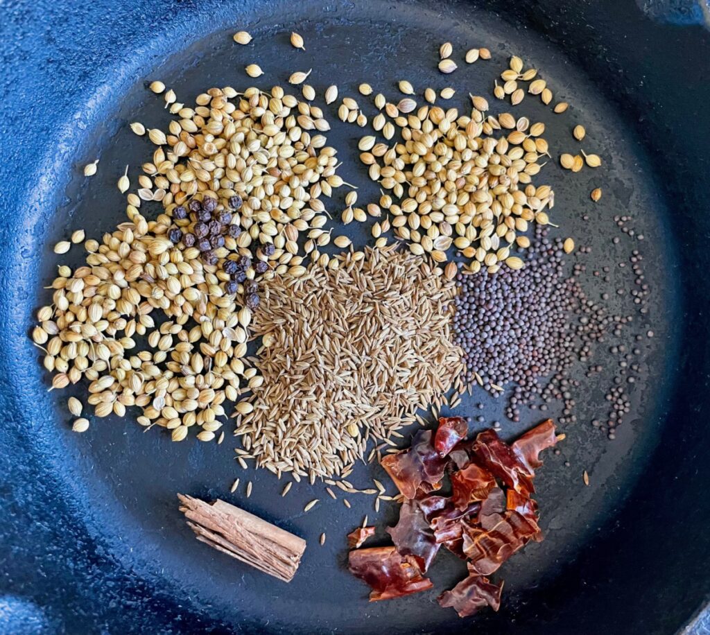 Whole spices for curry powder in a cast iron pan: coriander seeds, cumin seeds, brown mustard seeds, Ceylon cinnamon, red chilies, and black peppercorn.